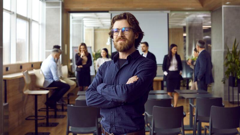 Fröhlicher, gutaussehender, bärtiger junger Mann in blauem Hemd und Brille, der nach einer Schulung im Büro steht und mit Überzeugung nach vorne blickt.